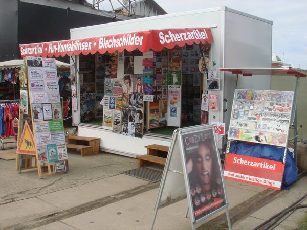 Verkaufsstand mit rotem Dachvorsprung, auf dem in Weiß „Scherzartikel • Blechschild • Fun-Kontaktlinsen“ steht. Vor dem Stand sind viele bunte Blechschilder und witzige Spruchtafeln dicht nebeneinander aufgestellt — z. B. Verkehrs- oder Retro-Motivschilder, Wortwitze, Markenlogos und Spruchtafeln. Der Stand ist ein kleiner mobiler Marktstand mit vielen Angeboten rund ums Schilder- und Scherzartikel-Sortiment.